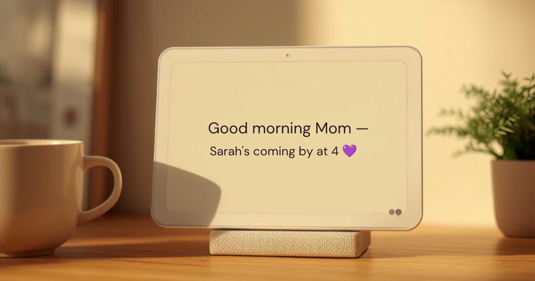 CareBoard display on a kitchen counter next to a coffee mug and a small plant, showing the message “Good morning Mom — Sarah's coming by at 4” in large type